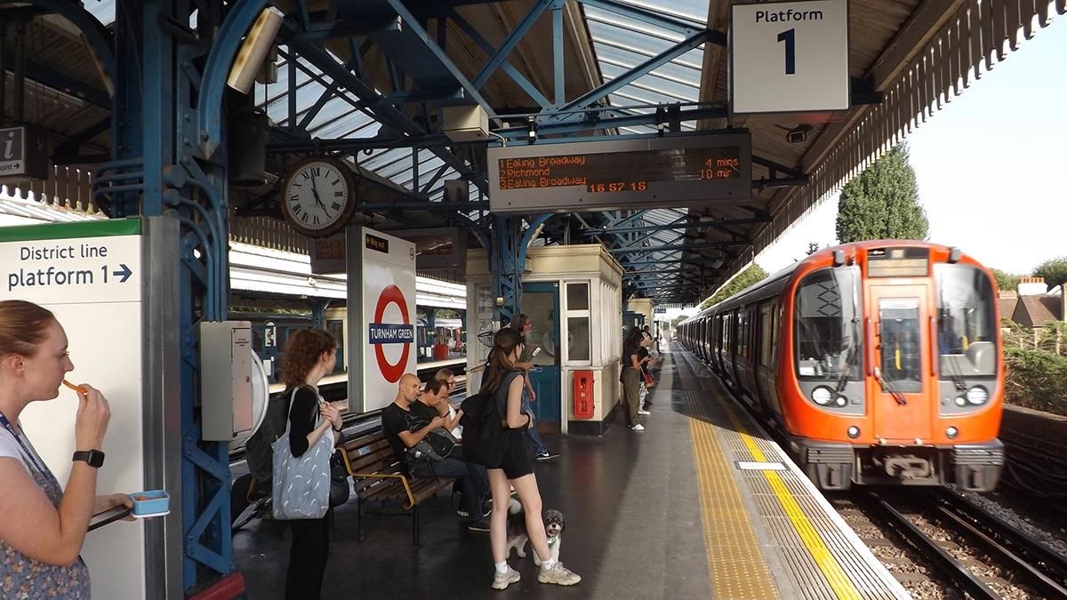 A busy surface level platform at Turnham Green station with commuters waiting as a train pulls into the station