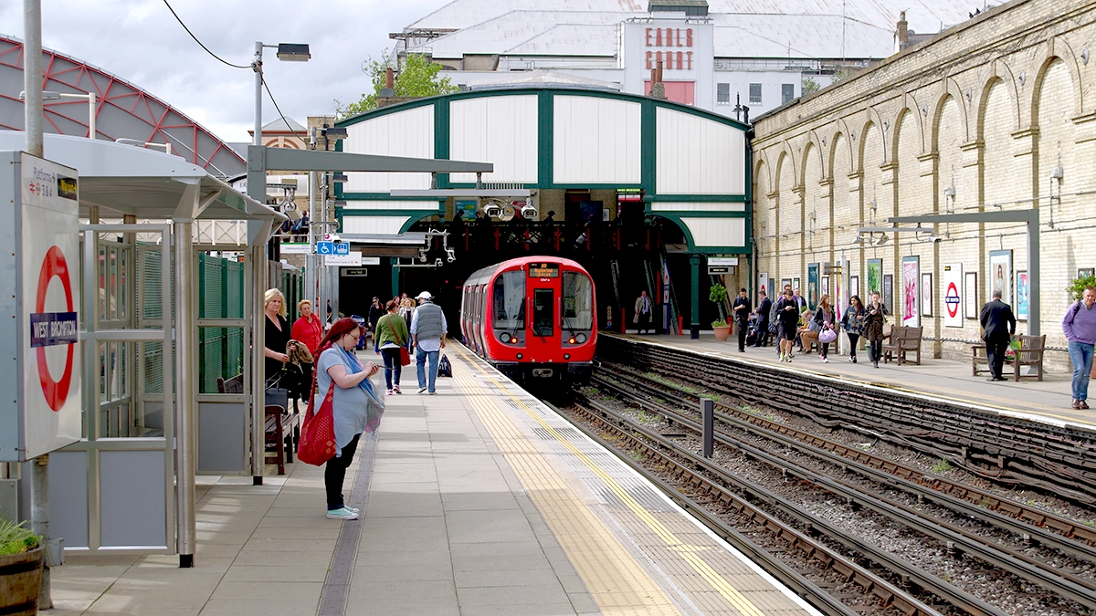 A tube train arrives at a busy platform at West Brompton with Earls Court Exhibition Centre in the background