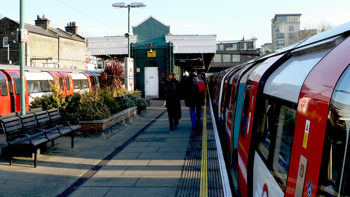 Commuters leave a tube train at Willesden Green station past the platform benches and planters